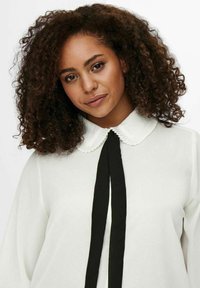 Blusa blanca con un collarete rizado, mangas largas y un lazo negro en el escote. Textura suave con un sutil brillo.