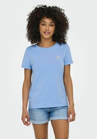 Camiseta de algodón azul claro con cuello redondo, mangas cortas y un pequeño diseño de corazón brillante en el lado izquierdo del pecho. Combinada con pantalones cortos de mezclilla.