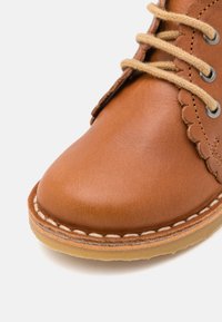 Chaussure en cuir marron avec un bout rond, des accents de design festonnés, des lacets beiges et un détail cousu le long de la semelle.