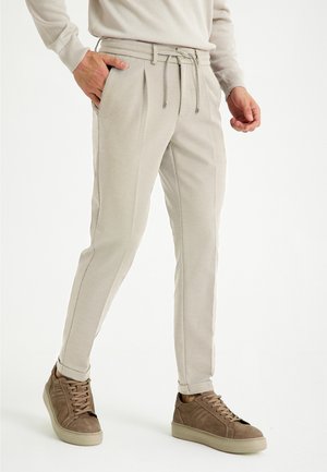 Beige joggerbroek gemaakt van geweven stof, met een trekkoord in de taille, twee zijzakken en contrasterende zijstrepen. Draag ze met lichtbruine sneakers.