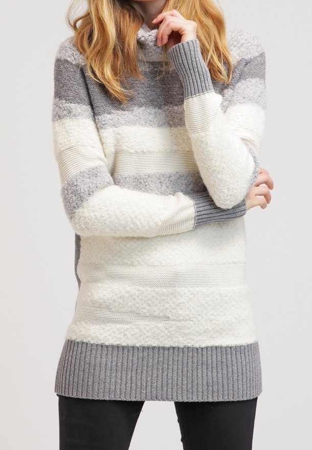 Femme portant un long pull en tricot rayé dans des tons de gris et blanc avec des motifs texturés, associé à un pantalon noir.