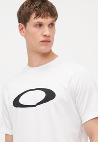 Weißes T-Shirt mit kurzen Ärmeln und einem großen schwarzen runden Logo auf der Vorderseite. Glattes Material mit moderner Passform und Rundhalsausschnitt.