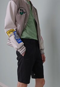 Veste varsity grise et blanche avec des patches, chemise verte en dessous et shorts noirs. La veste présente des détails brodés et des poignets rayés.