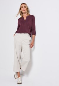 Femme aux cheveux blonds portant un haut violet à boutons, un pantalon large beige et des baskets beiges, adossée à un mur blanc.