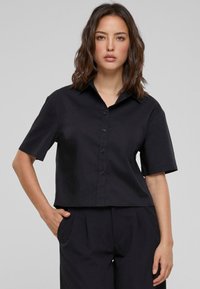 Camicetta nera a maniche corte con colletto, realizzata in tessuto liscio. Design cropped con orlo dritto, caratterizzata da una vestibilità ampia e senza fantasie.