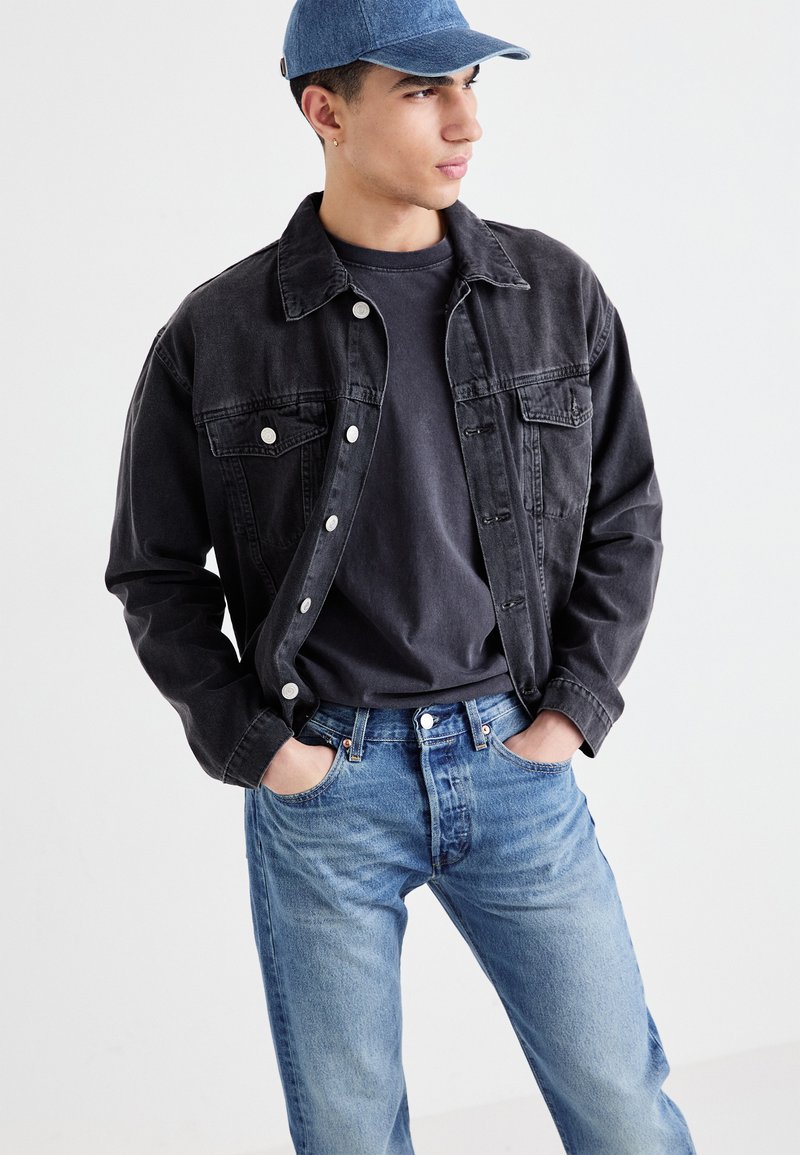 Schwarze Denimjacke mit zwei Brusttaschen, über einem dunklen Hemd getragen, kombiniert mit hellblauen Jeans. Akzente sind silberne Knöpfe und eine blaue Kappe.