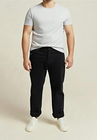 Homme portant un t-shirt blanc uni, un pantalon noir retroussé et des chaussures blanches sans lacets, debout devant un fond neutre.