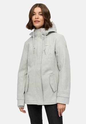 Femme portant un manteau d'hiver léger gris clair avec des poches avant, des cordons de serrage, et un pantalon noir sur un fond uni.