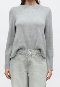 Personne portant un pull à manches longues gris clair et un jean gris délavé taille haute sur un fond uni.