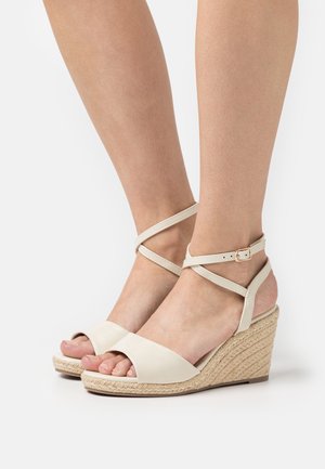 Sandalias tipo cuña en color beige con plataforma similar a paja, punta abierta y tiras cruzadas en los tobillos, que presentan un acabado de cuero suave y hardware mínimo.