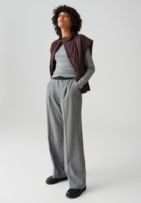 Femme aux cheveux bouclés portant un gilet marron sans manches sur une chemise grise à manches longues, un pantalon large gris et des chaussures noires, debout avec les mains dans les poches.