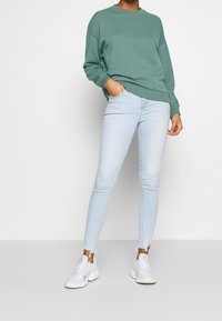 Sweatshirt vert avec des poignets et un ourlet côtelés, associé à un jean skinny bleu clair avec des chevilles déchirées et des chaussures de sport blanches.