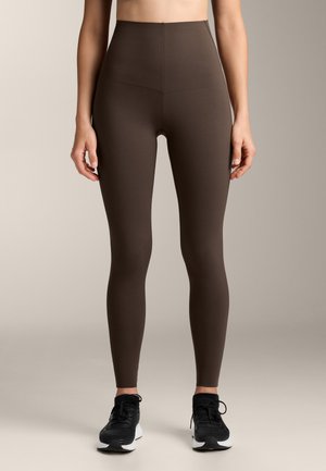 Colanți maro cu talie înaltă, din material elastic, având o textură netedă și un design ajustat, asortați cu adidași negri.