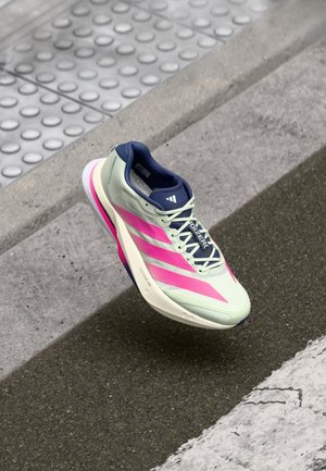 Hellgrüner und rosa Adidas Laufschuh schwebt über einem städtischen Gehweg in der Nähe taktiler Pflasterung und einer weißen Zebrastreifenlinie.