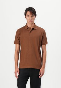 Hnědý polo tričko s proužky a límečkem, krátkým rukávem, kombinované s černými kalhotami. Měkká textura, přiléhavý design, bez viditelných vzorů či akcentů.