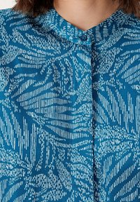 Blouse boutonnée en teal avec un motif abstrait blanc, un col rond et un tissu doux et texturé.