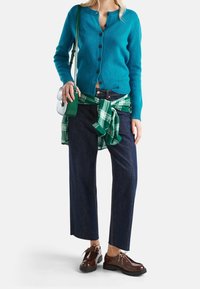 Turkos kofta med knappar, grön rutigt skjorta knuten vid midjan, mörka denim bredbyxor och bruna snörskor.