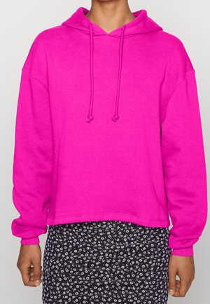 Personne portant un sweat à capuche rose vif avec cordons et une jupe noire à petit motif floral blanc.