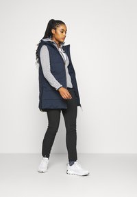 Marineblå puffervest med hætte, med lynlås fortil og to sidelommer, båret over en grå mønstret langærmet skjorte og sorte bukser. Hvide sneakers.