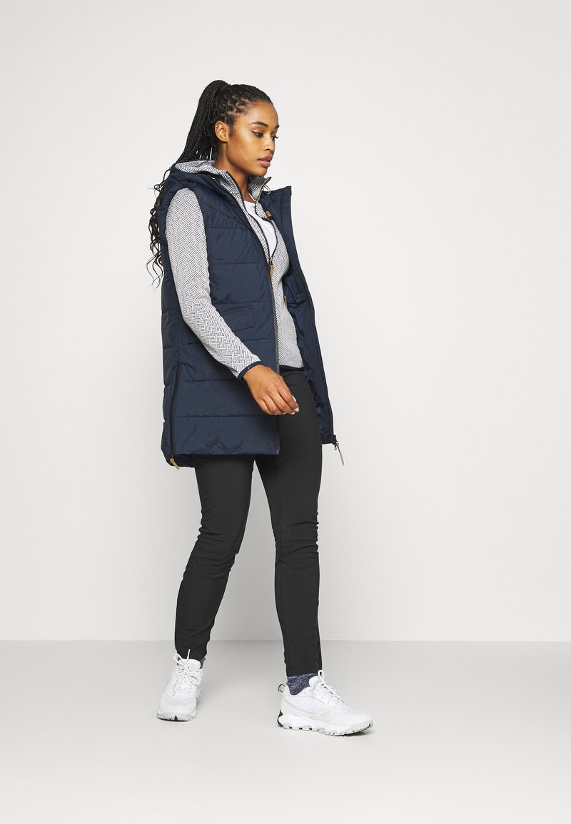 Gilet in piuma blu navy con cappuccio, dotato di zip frontale e due tasche laterali, indossato sopra una maglia a maniche lunghe grigia con motivo e pantaloni neri. Sneakers bianche.