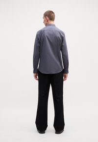 Ανδρικό πουκάμισο μακρυμάνικο σε σκούρο γκρι χρώμα, με κολάρο τύπου πόντ και κουμπωτούς καρπούς, φορεμένο με φαρδιά μαύρα παντελόνια, σε θέα από πίσω.