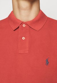 Červené polo tričko se zapnutými dvěma knoflíky, s malým modrým vyšitým logem polo hráče na hrudi.