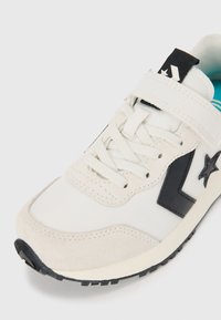 Sneakers blancs en matière suède et toile, avec des accents noirs, un logo en forme d'étoile, un design à lacets et une semelle en caoutchouc noir texturé.