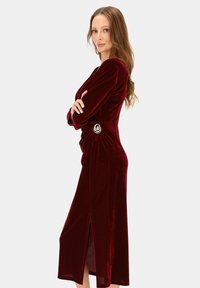 Bordowa aksamitna sukienka z długimi rękawami, bocznym rozporkiem oraz ozdobnym srebrnym akcentem w talii. Gładka faktura i dopasowany fason.