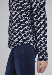 Navy blouse with a white swirling pattern, long sleeves, slightly rounded hem, paired with dark denim jeans. Smooth texture, relaxed fit.