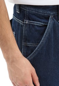 Jean en denim bleu foncé avec poches latérales, présentant des coutures contrastées et une coupe décontractée. Gros plan sur la taille et la position des mains.