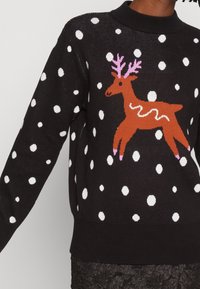 Pull en tricot noir avec des pois blancs et un motif de renne marron. Présente des bois roses et un détail en zigzag blanc sur le dos du renne.