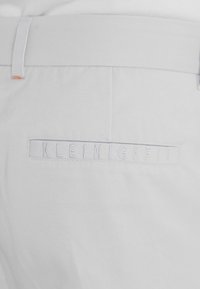 Pantalon blanc avec une poche arrière présentant un texte brodé subtil "KLEINIGKEIT" et une boucle de ceinture à la taille.