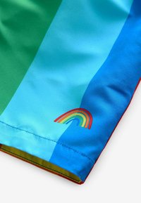 Shorts de bain colorés avec des panneaux bleus et verts, texture lisse et un petit graphisme arc-en-ciel brodé sur la jambe. Accents rouges.