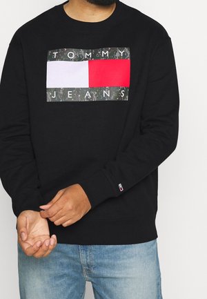 Uomo che indossa una felpa nera con un grande logo Tommy Jeans composto da blocchi bianchi e rossi, abbinata a jeans azzurri, con le mani incrociate.