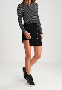 Gonna nera con dettagli di bottoni argentati, abbinata a una maglietta a righe bianco e nera a maniche lunghe. Il modello indossa sneakers alte nere.