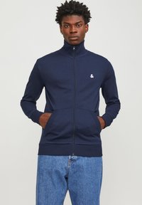 Navy zip majica s visokim ovratnikom, prednjim džepovima, rebrastim manžetama i bijelim logotipom. Izrađena je od mekog, glatkog materijala.