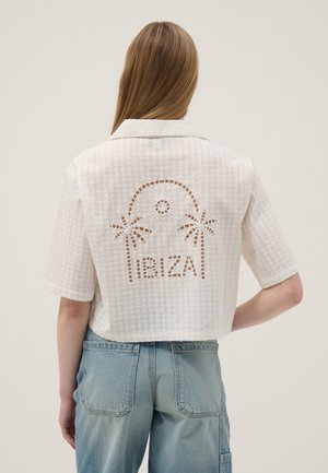 Mujer con cabello largo que lleva una camiseta blanca de manga corta con el diseño recortado de "IBIZA" y una palmera en la espalda, y jeans azul claro.
