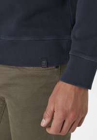 Marineblaue Sweatshirt mit strukturiertem Stoff, gerippten Bündchen und einem kleinen Etikett mit "P." an der unteren Seite, kombiniert mit khakifarbene Hosen.