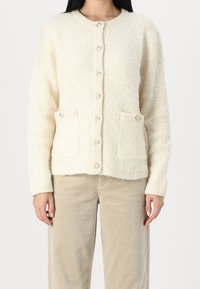 Krämfärgad cardigan i texturerat, fluffigt material; har rund hals, framknappar och två framfickor.