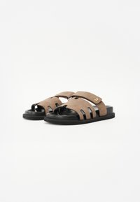 Brune suede sandaler med sorte såler, der har udskårne mønstre og en bred strop over foden. Gulddetalje på stroppen.