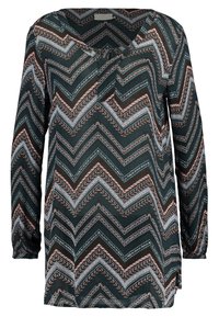 Blouse à manches longues vert foncé, avec un motif chevron en marron, rose et bleu pâle. Encolure en V, coupe décontractée et fentes latérales.