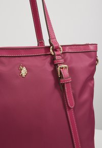 Ciemnoróżowa torba na ramię z regulowanym paskiem, złotą klamrą i małym złotym emblematem przedstawiającym konia i jeźdźca na jednolitym tle.