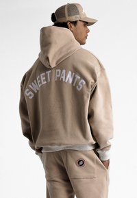 Hoodie beige avec "SWEET PANTS" brodé au dos. Tissu doux, coupe décontractée, et pantalon assorti avec un logo en patch sur le côté.