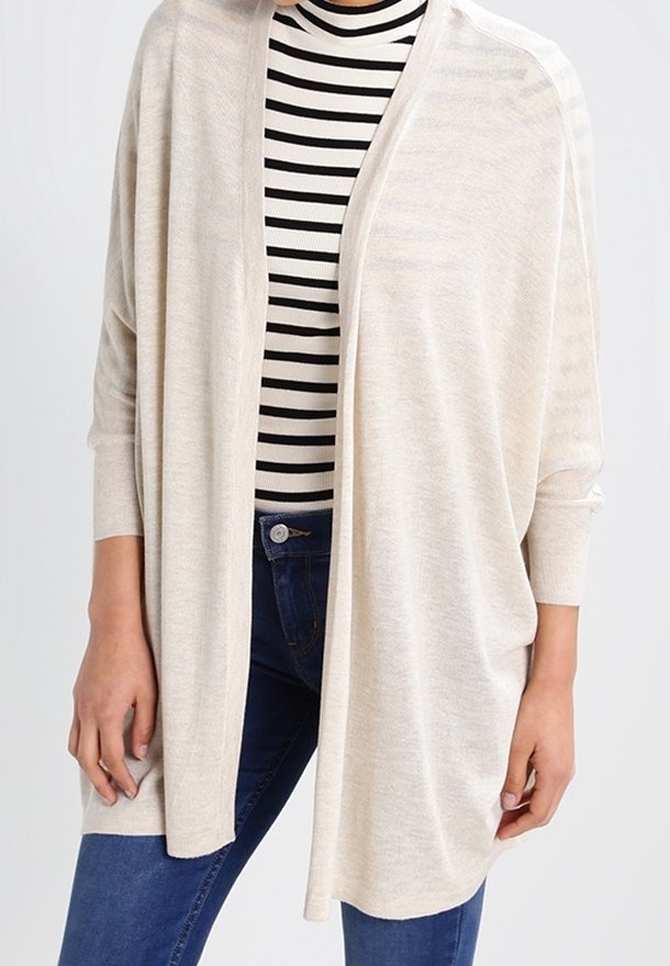 Gilet beige clair ouvert avec une coupe ample, des manches longues et une texture douce, superposé sur un haut rayé noir et blanc.