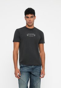Hombre con camiseta de manga corta negra "GUESS" y vaqueros azules, de pie contra un fondo blanco, mirando hacia adelante.