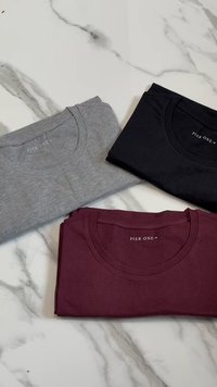 Tre T-shirt piegate in grigio, nero e bordeaux. Ognuna presenta un colletto rotondo e una consistenza morbida simile al cotone. Etichetta del marchio visibile.