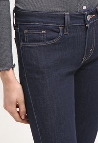 Jeans de mezclilla azul oscuro, con cinco bolsillos, costuras visibles en naranja contrastante y un ajuste ceñido en la cintura y el muslo.