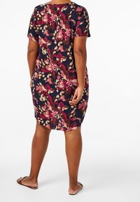 Kvinna som står barfota och bär en knälång, kortärmad klänning med ett abstrakt blommönster i marinblått, rosa, brunt och beige.