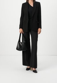 Schwarzer taillierter Blazer über einem schwarzen Oberteil mit passender Hose; kombiniert mit einer schwarzen Umhängetasche und spitzen schwarzen High Heels.