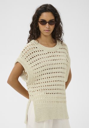 Kiharalukkade ja tumeprillidega naine, kes kannab varrukateta kreemja tooni kootud kampsunit külgede lõhikute ja valge rõivastuse peal.